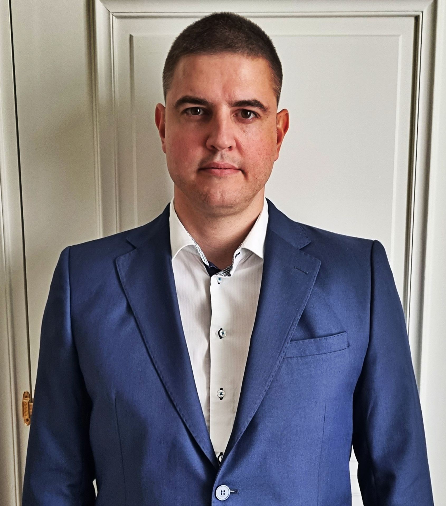 Individual wearing a blue blazer and white dress shirt, standing in front of a white door.
